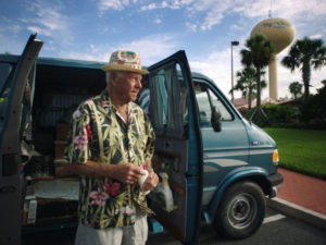 Dennis, wearing a floral Hawaiian shirt that matches with his hat, stands in front of his trusty van.