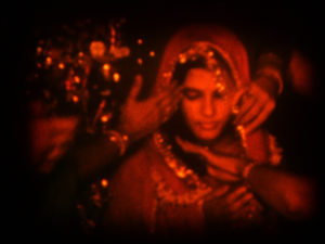 The image, a still from A Night of Knowing Nothing, is in a deep red hue. An Indian woman is dressed in clothing seemingly being prepared for a special occasion..