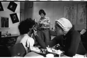 In a Black and White still from Dope is Death, a Black woman, who is wearing a head wrap, performs acupuncture on another Black woman. The woman performing the acupuncture looks focused, and there is a white man in the middle background of the image working on something else.
