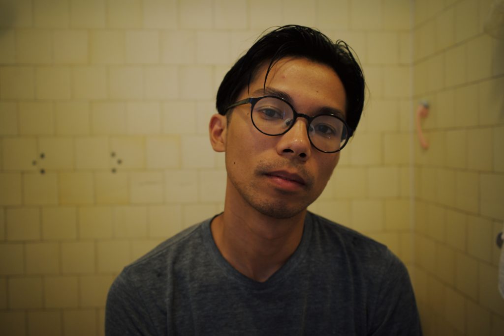 The image is portrait of filmmaker Miko Revereza. He is a Filipino man with dark hair, and he is wearing glasses and Black shirt in this portrait.