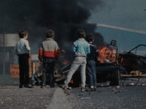 Four young boys stand on the pavement, looking at burning debris.