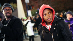 In a still from the film, Unapologetic, Bella stands at a protest, facing partially out of frame. She is wearing a black jacket and red hoodie, and another man stands to the left of her, facing a similar direction.