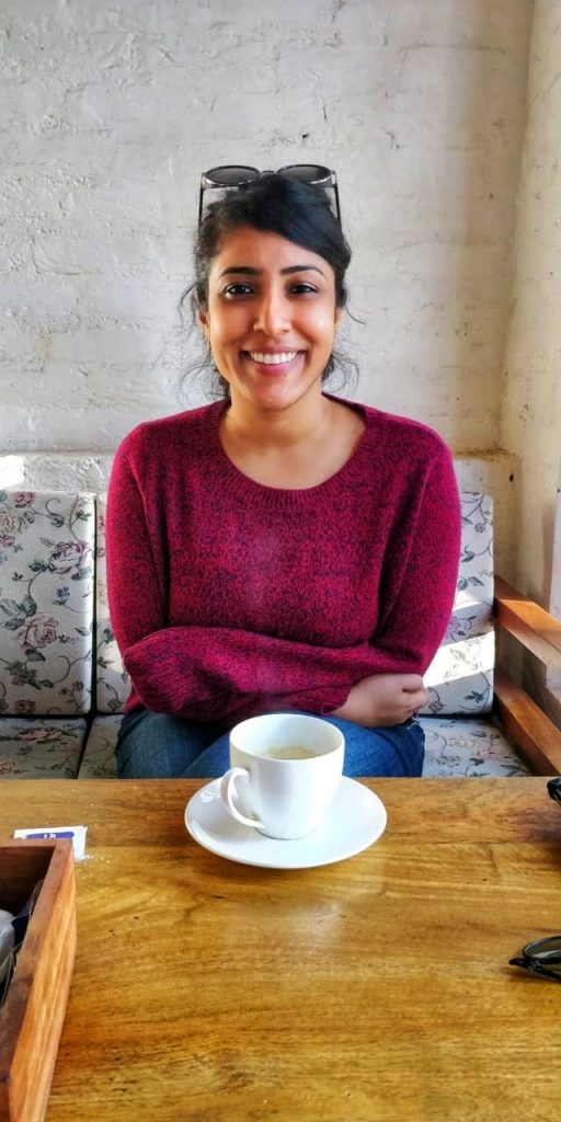 The image is a portait of filmmaker Rintu Thomas. She is an Indian woman with dark hair, and sunglasses on top of her head. She is smiling brightly at the camera as she sits at a table with a white coffee cop sitting in front of her.