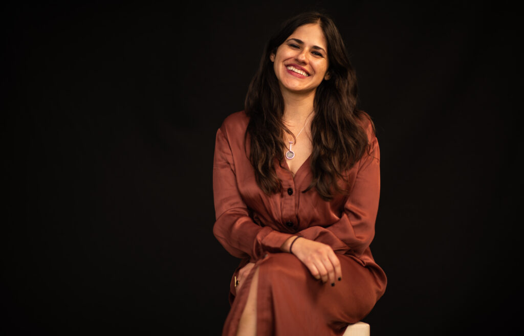 The image is a portrait of filmmaker Iliana Sosa. She is a Mexican-American woman, and she's sitting in the portrait. She has long dark hair and is wearing silver jewelry and a brown dress as she smiles warmly to the camera.