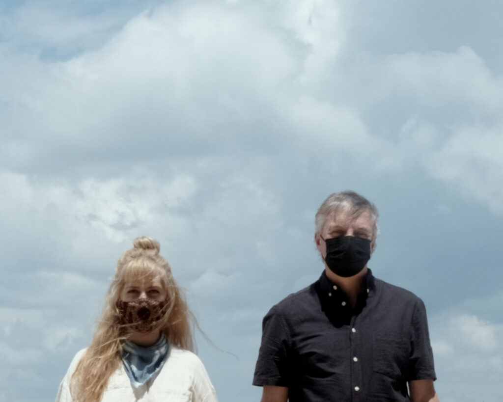 The image is a portrait of the filmmakers Erin and Travis Wilkerson. They're standing in front of a group of clouds. Erin is a white woman with long blonde hair and she is wearing a bandana as a face covering. Travis, on the right, is a white man with salt and pepper hair. He is wearing a black mask on his face.