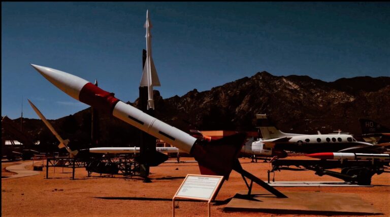 In this image, a white rocket can be seen in the desert. Mountains are in the distance behind them.