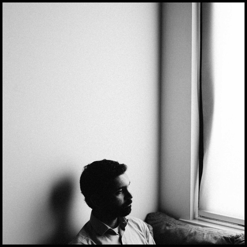 The image is a black and white portrait of the filmmaker, Saeed Taji Farouky. He is a Palestinian-British man, and he is sitting in front of a window with natural light coming in. The filmmaker is sitting next to the window and he's looking towards the window away from the camera.