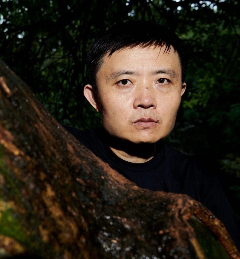 The image is a portrait of filmmaker FAN Jian. He is a Chinese man with black hair. He is staring forward in the image as he stands next to a tree that foregrounds the image.