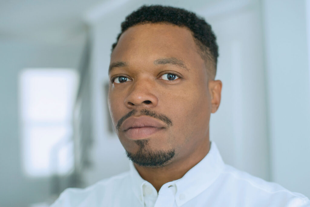 A portrait of filmmaker Jon-Sesrie Goff. He is a Black with low cut hair and a beard goatee. He is looking calmly at the camera and is wearing a white button down.