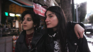 In an image from the film, Sirens, Lilas and Shery are walking down a street with their arms around each other. Lilas arm is on Shery's shoulders. They are both Lebanese women with dark hair, and are wearing dark, largely black clothing.