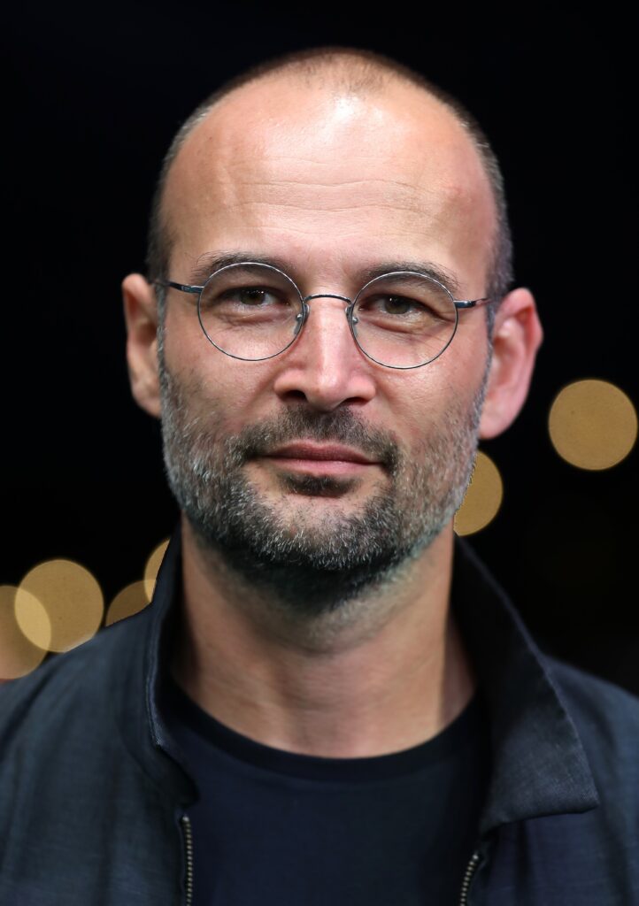The image is a portrait of the German-Romanian filmmaker, Alexander Nanau. filmmaker Alexander Nanau. He has a low-cut haircut, and he is wearing classes. Flashes of light can be seen behind him.