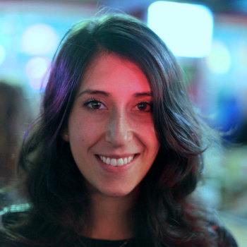 The image is a headshot of filmmaker Rita Baghdadi. She is a Moroccan-American woman with brown hair, and there is a red tint of light illuminating her face. She is smiling kindly at the camera.